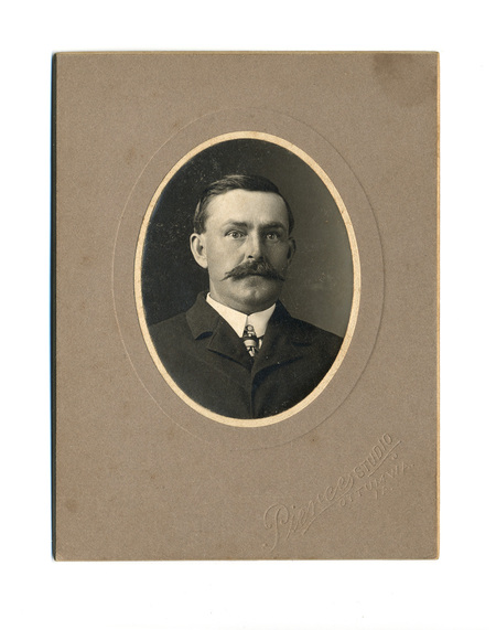 Retrato de estudio de un hombre adulto capturado a principios del siglo XX. El sujeto luce un bigote tupido tipo manubrio, viste traje oscuro de solapa ancha, camisa blanca con cuello rígido y corbata con un alfiler ornamental. La composición es un busto frontal dentro de un marco ovalado sobre un soporte rígido.