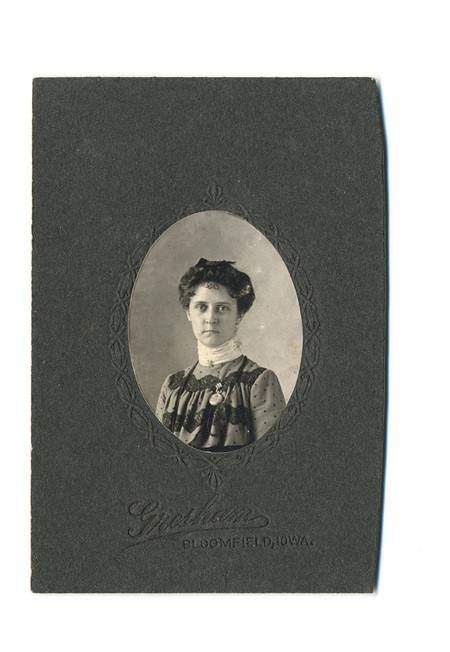 Retrato de estudio en formato busto de una mujer joven. Viste una prenda de tonos oscuros con aplicaciones de encaje y una blusa de cuello alto blanco. Porta un pequeño dije o medalla en el pecho y su cabello está peinado hacia arriba siguiendo la moda de la época. La fotografía está contenida en un óvalo decorativo sobre un soporte rígido.