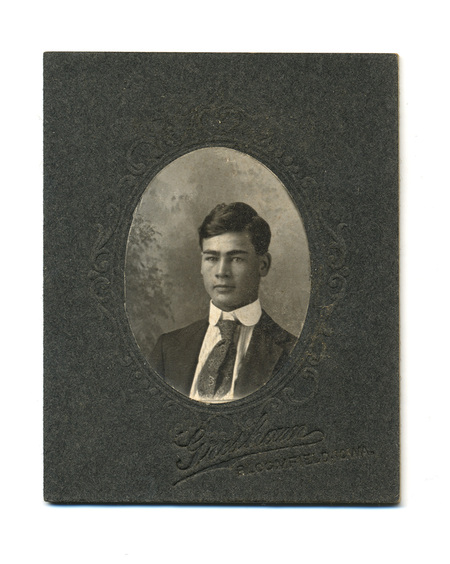 Retrato de estudio de un hombre joven de cabello oscuro y rasgos marcados, capturado en una pose de tres cuartos. Viste un traje formal con una camisa de cuello rígido y una corbata de seda con intrincados motivos ornamentales. La fotografía está contenida en un óvalo impreso sobre una montura de cartón grisáceo decorada con filigranas en relieve, característica de los estudios fotográficos comerciales estadounidenses de la época.