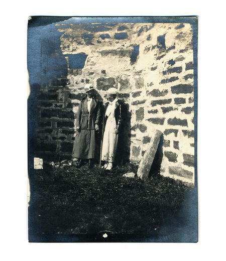 Fotografía de principios del siglo XX que captura a dos mujeres posando frente a los restos de una edificación de piedra, probablemente una estructura colonial en ruinas. Ambas visten abrigos largos y sombreros típicos de la época posrevolucionaria. La escena muestra una composición estática donde destaca el contraste entre la vestimenta urbana y el entorno rústico de la construcción deteriorada.