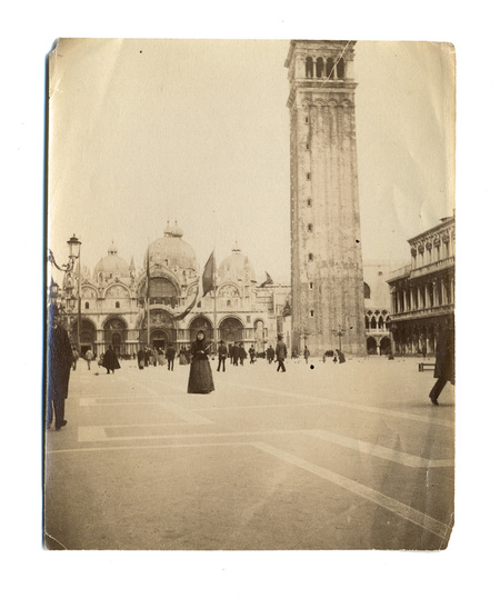 Captura urbana de finales del siglo XIX que muestra la Plaza de San Marcos en Venecia. Al fondo se aprecia la fachada de la Basílica y a la derecha el Campanile. La escena registra el tránsito de personas con vestimentas de época, resaltando una mujer en el centro con traje oscuro y sombrero.