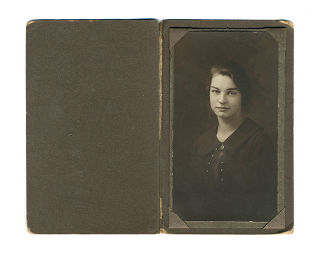 Retrato de estudio de una mujer joven de mirada frontal y expresión serena, peinada con un estilo ondulado característico de las décadas de 1920 o 1930. Viste una prenda oscura de cuello amplio con detalles ornamentales de pequeños botones o apliques en el frente. La imagen está montada en una carpeta protectora de cartón oscuro.