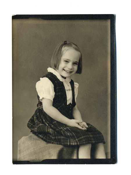 Retrato de estudio de una niña pequeña, capturado aproximadamente a mediados del siglo XX en México. La infante aparece sentada de perfil, girando el rostro hacia la cámara con una sonrisa expresiva. Viste un atuendo escolar o de diario formal consistente en un jumper de cuadros sobre una blusa blanca, reflejando las normas de vestimenta infantil urbana de la época.