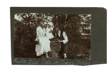 Retrato familiar al aire libre capturado en 1919. Una mujer en vestido blanco de encaje y un infante posan junto a un joven vestido formalmente con chaleco y moño. La escena, situada frente a follaje denso, incluye un perro en la esquina inferior derecha y una inscripción manuscrita identificativa en el margen inferior del soporte.