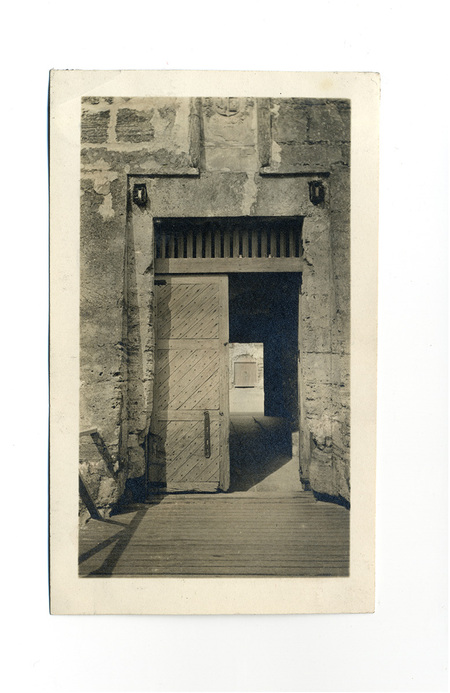 Vista de la entrada principal a una fortificación colonial española, capturada desde el puente de acceso. Se observa una puerta de madera reforzada con herrajes, situada bajo un arco adintelado de piedra labrada que ostenta un escudo heráldico. La perspectiva conduce la mirada hacia el interior del recinto fortificado a través de un pasillo sombreado.