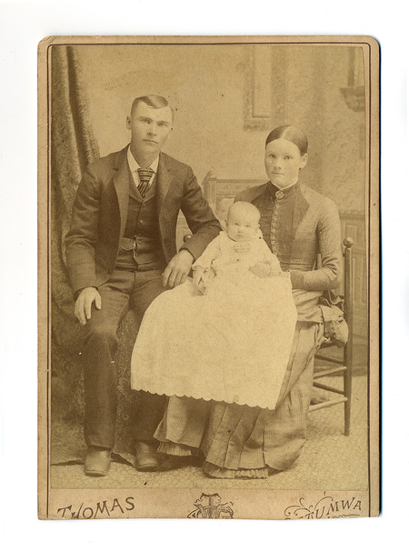 Retrato de estudio de una familia joven compuesto por un hombre, una mujer y un infante. El hombre posa sentado con un traje oscuro y cadena de reloj visible; la mujer viste una prenda de tonos oscuros con detalles abotonados. En el centro, un bebé luce un extenso ropón blanco de encaje. La composición incluye un telón de fondo con cortinajes y una silla de madera torneada.