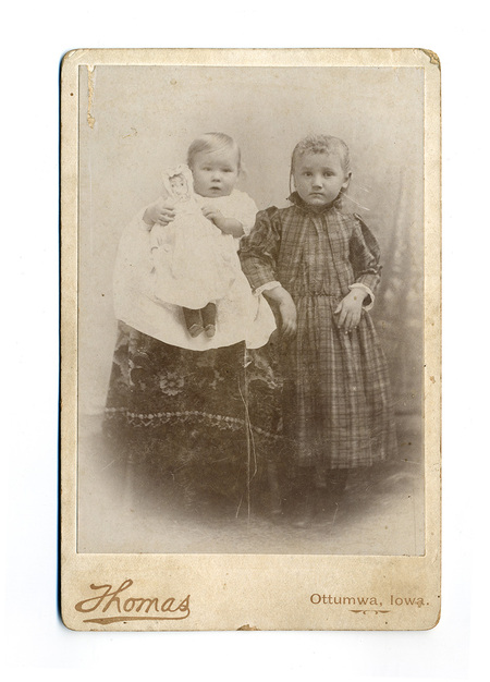 Retrato de estudio de dos infantes a finales del siglo XIX. Un ni&ntilde;o de corta edad se encuentra sentado sobre un pedestal cubierto con una alfombra de motivos florales, sosteniendo una mu&ntilde;eca de trapo. A su lado, una ni&ntilde;a mayor permanece de pie vistiendo un vestido de cuadros oscuros. Ambos presentan expresiones serias, t&iacute;picas de los largos tiempos de exposici&oacute;n de la &eacute;poca.