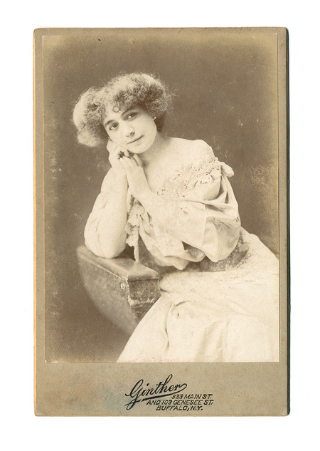 Retrato de estudio en plano medio de una mujer joven con cabello rizado y voluminoso. Viste un elegante traje de tonalidad clara con profusos detalles de encaje en el escote y las mangas. La figura posa de tres cuartos, con el brazo izquierdo apoyado sobre un mueble de estudio y las manos juntas cerca del rostro en una actitud contemplativa.