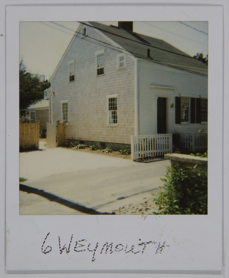 Vista exterior de una residencia de estilo colonial tradicional con fachada de tejas de madera y carpintería blanca. La escena muestra un cerco de madera tipo 'picket fence' y una entrada vehicular de grava. La imagen, capturada con luz solar directa, presenta una inscripción manuscrita en el borde inferior que identifica la ubicación como '6 Weymouth'.