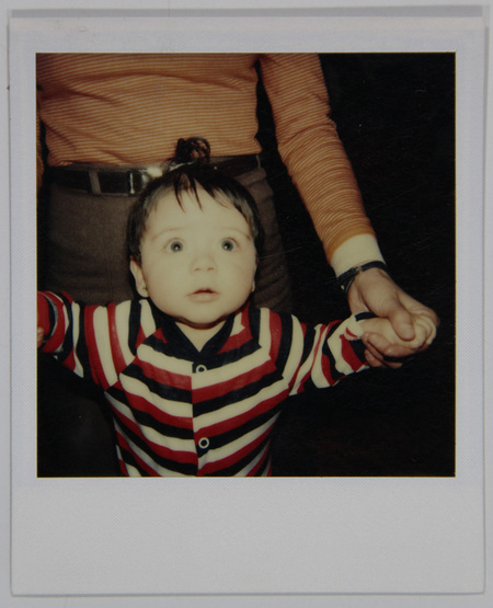 Retrato doméstico de un infante con expresión de asombro, vestido con una prenda de rayas horizontales en color rojo, negro y blanco. El menor es sostenido por las manos de una persona adulta, de la cual solo se observa el torso con una camisa de rayas ocres y pantalones oscuros. La escena captura un momento cotidiano de crianza típico de la clase media urbana.