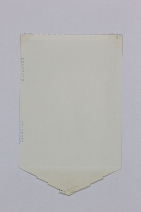 Vista posterior de una fotografía instantánea Polaroid. Se observa un soporte de papel color crema con la marca del fabricante y un número de serie estampados lateralmente en tinta azul. El diseño incluye bordes troquelados y una pestaña en forma de flecha en el extremo inferior, elementos funcionales del antiguo proceso de revelado por difusión-transferencia manual.