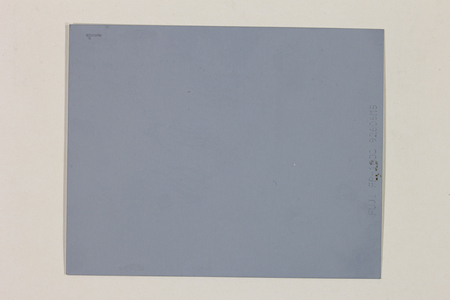 La imagen muestra el reverso de una película instantánea de formato profesional. No presenta una escena fotográfica visible, sino una superficie plana y uniforme de color gris azulado. En el borde derecho se observa un sello técnico de fábrica con el modelo del producto, y en la esquina superior izquierda se aprecia un número '1' escrito manualmente.
