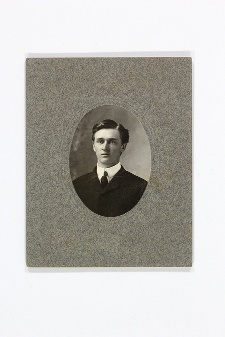 Retrato de estudio de un hombre joven en un encuadre de busto con corte ovalado. El sujeto presenta cabello corto peinado hacia atrás y viste un saco obscuro sobre una camisa blanca con cuello alto rígido y corbata obscura. El retrato está montado sobre una cartulina gris de grano grueso con relieve tipográfico.