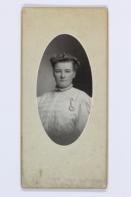 Retrato de estudio de una mujer joven capturado en un formato vertical. La figura aparece de medio cuerpo, luciendo una blusa blanca con detalles de bordado y un broche circular prendido al pecho. Su cabello está peinado con ondas hacia atrás, y mantiene una expresión formal frente a un fondo neutro degradado.