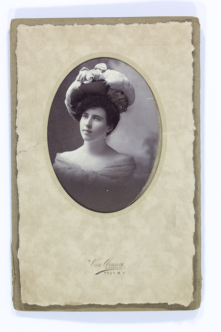 Retrato de estudio de una mujer joven capturada en un formato ovalado. La modelo luce un peinado elevado y un sombrero de gran escala adornado con motivos textiles, complementado con un vestido de hombros descubiertos. Su pose es de tres cuartos, mirando ligeramente hacia arriba en una actitud contemplativa típica de la época.