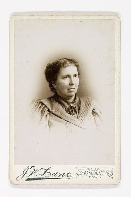 Retrato de estudio de una mujer adulta de tez clara con el cabello recogido. Viste una blusa de cuello alto con aplicaciones decorativas lineales y hombros ligeramente estructurados, moda típica de finales del siglo XIX. La composición utiliza un efecto de viñeta que difumina el cuerpo hacia la base del soporte, centrando la atención en el rostro de la retratada.