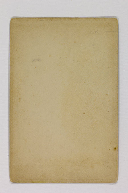 Esta imagen muestra el reverso o verso de una fotografía histórica. Se aprecia un soporte de cartón rígido con pátina de envejecimiento, esquinas redondeadas y diversas manchas de humedad y oxidación (foxing). En la esquina superior derecha se observa una breve anotación manuscrita a lápiz con el número '2'.