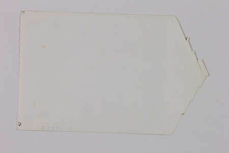 Reverso de una impresión fotográfica de mediados del siglo XX que muestra un borde lateral con acabado festoneado y diversas marcas de catalogación de laboratorio. La pieza presenta una forma irregular y signos de deterioro natural como manchas de oxidación y amarillamiento del papel, conservando códigos numéricos impresos que servían para la identificación industrial de la copia.