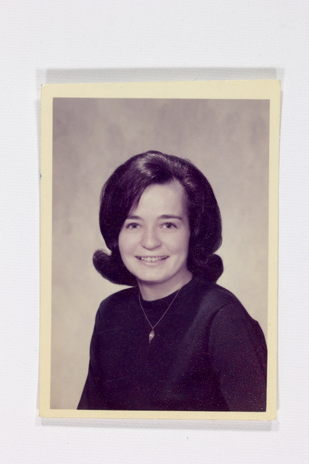 Retrato de estudio de una mujer joven, datado aproximadamente en la decada de 1960. La figura luce un peinado estilo 'flip' con puntas hacia afuera y volumen superior, caracteristico de la moda de mediados del siglo XX. Viste una blusa oscura de cuello redondo y un collar con dije alargado frente a un fondo neutro difuminado.