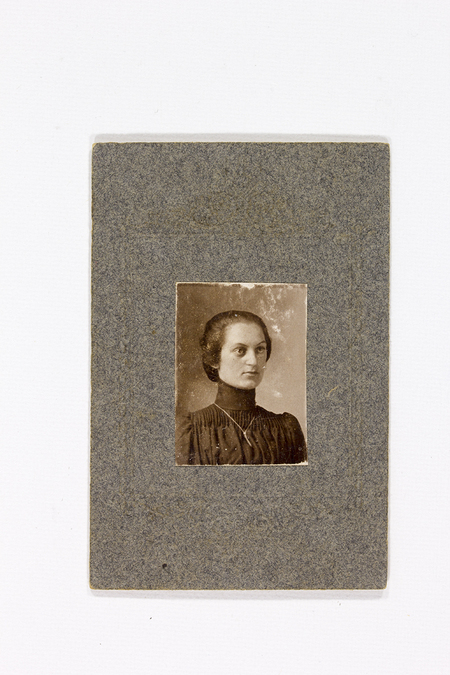Retrato de estudio de una mujer joven en una toma de busto. La retratada viste una blusa o vestido oscuro de cuello alto y pechera con finos pliegues verticales, complementado con una cadena delgada. Su cabello está recogido de manera sobria. La imagen está montada sobre un cartón grisáceo de grandes dimensiones con un marco decorativo en relieve.