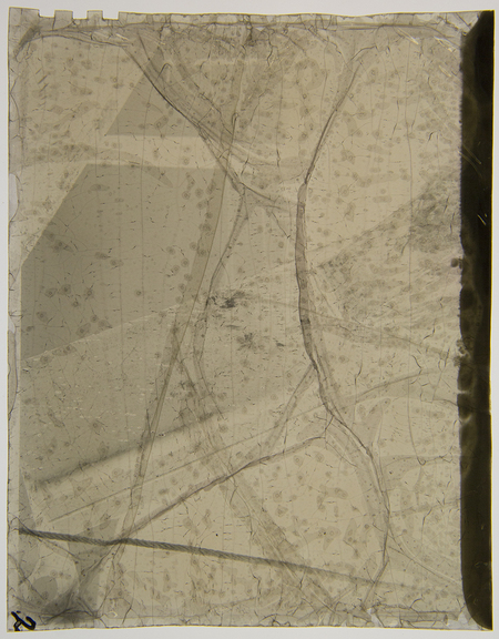 Imagen histórica en estado avanzado de degradación física que muestra fragmentos de una escena documental del periodo revolucionario mexicano. La descomposición química del soporte ha fracturado la emulsión, creando un entramado de grietas y pliegues que oscurecen los detalles originales de la composición, dejando apenas visibles formas que sugieren estructuras arquitectónicas o figuras humanas en un entorno urbano.
