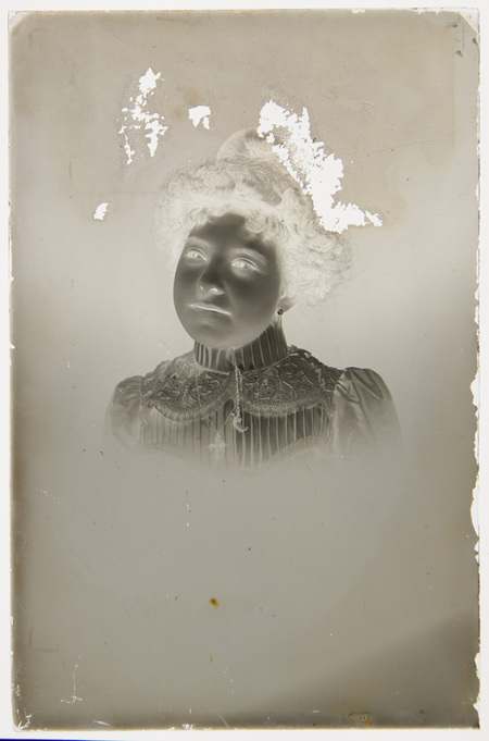 Retrato de estudio de una mujer joven con vestimenta formal de finales del siglo XIX o inicios del XX. La figura porta una blusa de cuello alto decorada con encaje intrincado y joyería que incluye una cruz y un dije de medialuna. Su peinado recogido y el estilo de la prenda sugieren una pertenencia a la clase media o alta urbana de la época.