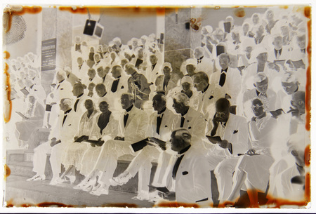 Imagen en negativo que captura a una multitud sentada en gradas durante una ceremonia oficial. Los asistentes, vestidos con ropa formal de gala, incluyen hombres de edad avanzada con trajes oscuros y mujeres con vestidos de media gala y sombreros. Un cartel informativo en una columna a la izquierda anuncia un evento conmemorativo relacionado con países escandinavos, sugiriendo una reunión diplomática o cultural internacional.