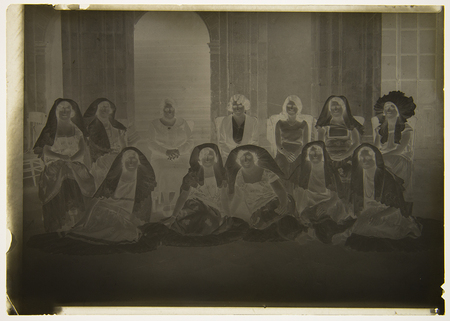 Un retrato grupal de doce mujeres posando en un espacio interior con arquería de piedra. Las mujeres están dispuestas en dos filas, algunas sentadas en sillas y otras en el suelo. Visten una mezcla de indumentaria regional mexicana, destacando mantillas de encaje y tocados tradicionales del Istmo de Tehuantepec, junto con vestidos de estilo contemporáneo de la primera mitad del siglo XX.