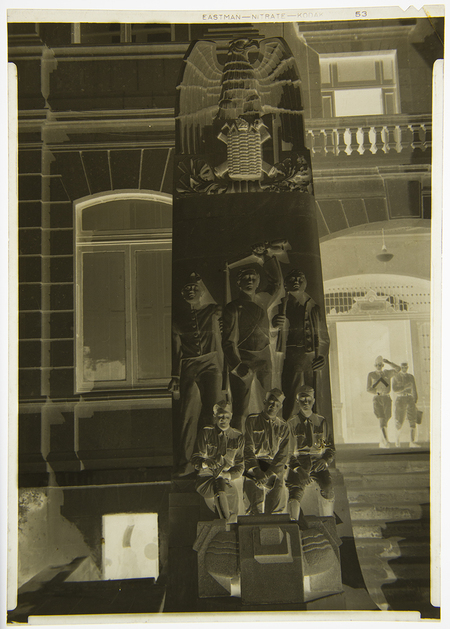 Fotografía en negativo que muestra un monumento conmemorativo de estilo modernista con relieves de cadetes y el escudo nacional mexicano en la parte superior. La escena se desarrolla frente a un edificio de arquitectura institucional o académica, donde se aprecia a dos individuos portando uniformes militares que caminan hacia un pórtico iluminado al fondo.