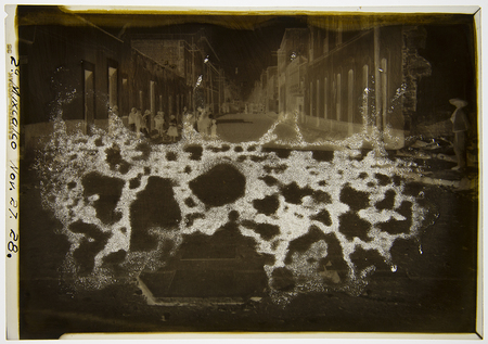 Escena urbana capturada en una calle de arquitectura colonial en México a finales de 1928. En la composición se aprecian diversas figuras humanas, incluyendo hombres con sombreros de ala ancha y mujeres con rebozos, situados frente a fachadas con puertas altas y molduras clásicas. La imagen documenta la atmósfera social y el entorno construido durante el periodo del Maximato.