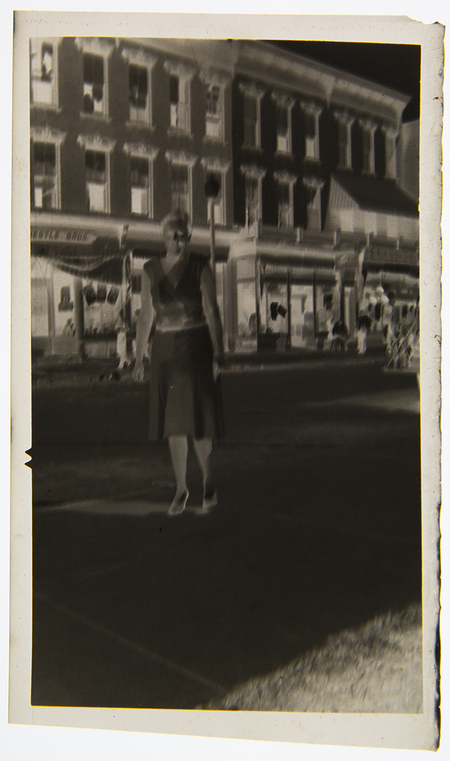 Retrato de un hombre joven de pie frente a un automóvil descapotable de mediados de la década de 1930. El individuo viste un suéter tipo cárdigan oscuro y pantalones de tono claro. La escena se desarrolla en una calle urbana con edificios de ladrillo al fondo y un árbol sin hojas, lo que sugiere una estación invernal o de otoño.