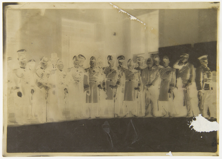 Retrato grupal de aproximadamente quince figuras masculinas ataviadas con uniformes militares o diplomáticos de gala, alineadas en un recinto oficial. Los hombres visten guerreras oscuras con botones metálicos, charreteras, condecoraciones y portan espadines ceremoniales; algunos usan bicornios y otros gorras de plato. La imagen muestra una severa deformación física y química en su soporte.