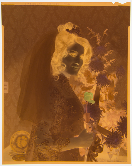 Retrato de estudio de una persona de género femenino con un peinado alto y elaborado, adornado con flores y un velo traslúcido. La figura se encuentra frente a un fondo de papel tapiz con un patrón floral de tipo damasco. La composición y el estilo sugieren un retrato formal, probablemente de boda o de una ceremonia social significativa de mediados del siglo XX.