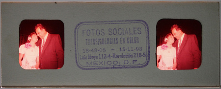 Vista estereoscópica nocturna que muestra un automóvil Volkswagen Sedán decorado con pintura blanca. En el costado se lee 'DE COLORES Nº 25 TAMPICO', lo que sugiere la participación en un evento religioso o caravana social. Dos figuras masculinas vestidas de traje formal oscuro se encuentran junto al vehículo, una de ellas apoyada en la parte trasera, frente a una estructura arquitectónica moderna con celosías.